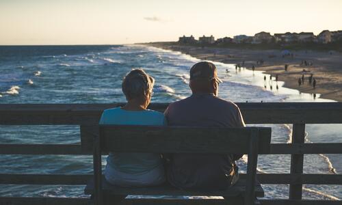 An elderly couple sitting on a bench from the pier facing the ocean