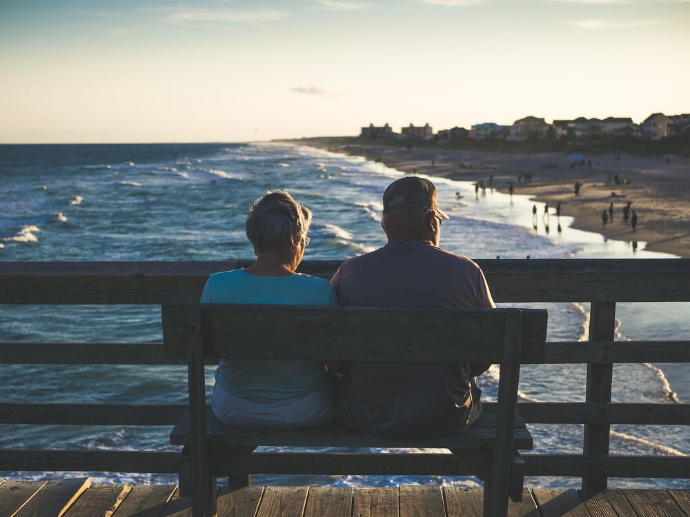 An elderly couple sitting on a bench from the pier facing the ocean